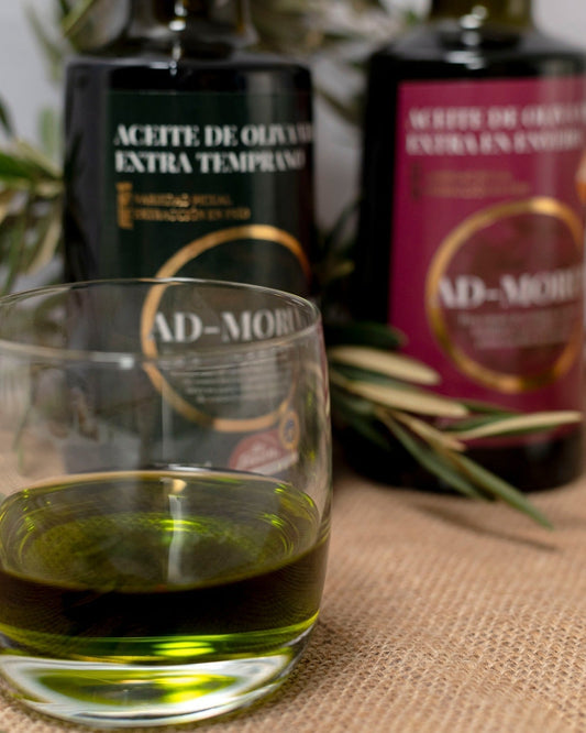 Vaso de aceite de oliva virgen extra temprano (AOVE Temprano) Ad-Morum junto con botellas de Ad-Morum al fondo.